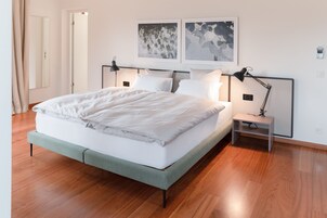 Una cama bien hecha con ropa blanca, una mesita de noche con una lámpara y dos cuadros en la pared.