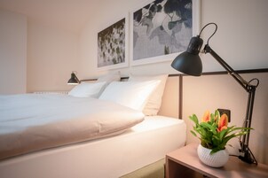 A neatly made bed with white linens, a bedside lamp, and a small table with a potted plant.