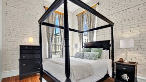 A four-poster canopy bed with a white bedspread and green pillows, a nightstand with a lamp, and a dresser with drawers. - Broughton Retreat in the Heart of Downtown! (Savannah)