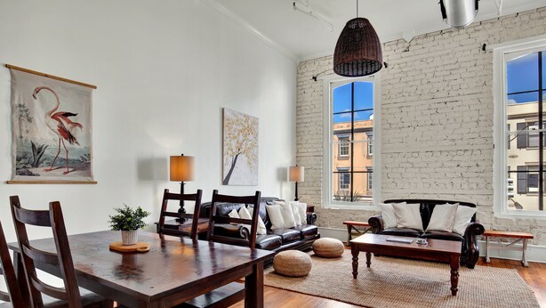 A spacious living room with a wooden dining table, leather sofa, and a large window with a view of buildings. - Broughton Retreat in the Heart of Downtown! (Savannah)
