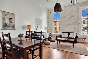A spacious living room with a wooden dining table, leather sofa, and a large window with a view of buildings. - Broughton Retreat in the Heart of Downtown! (Savannah)