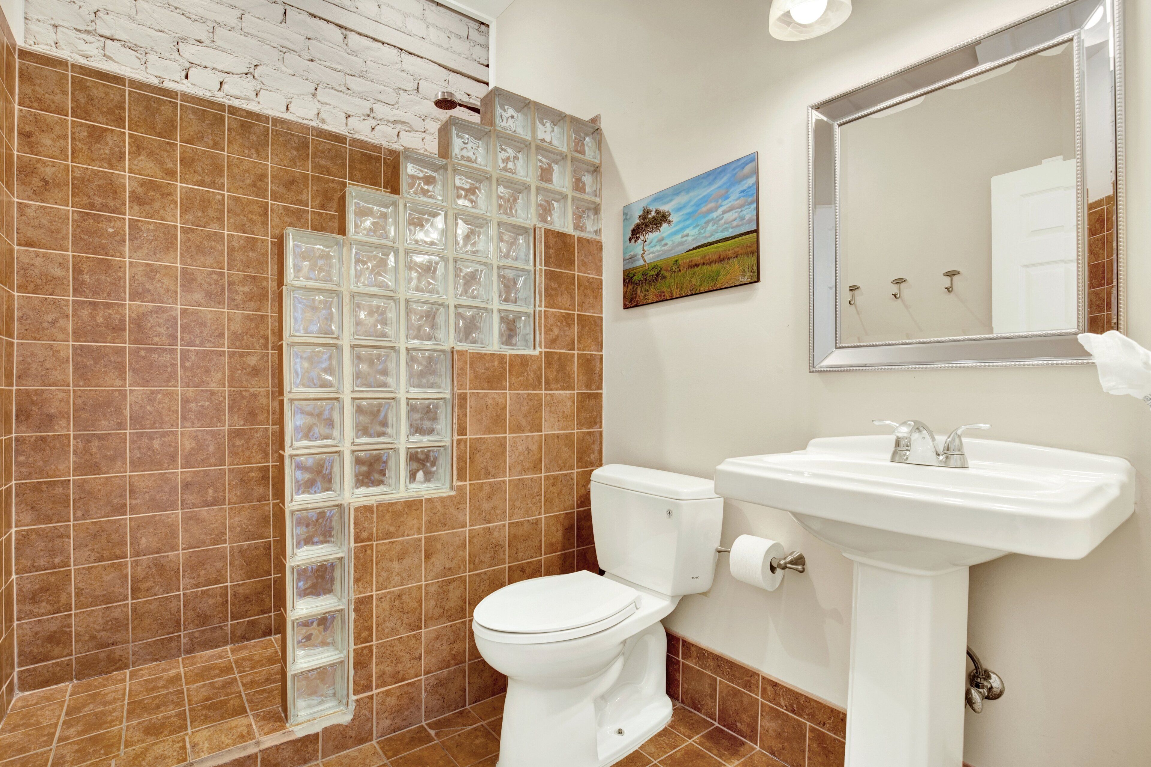 A bathroom with a white toilet, a sink, and a mirror. There is a window with a view of a field and a tree. The walls are tiled in brown and white.