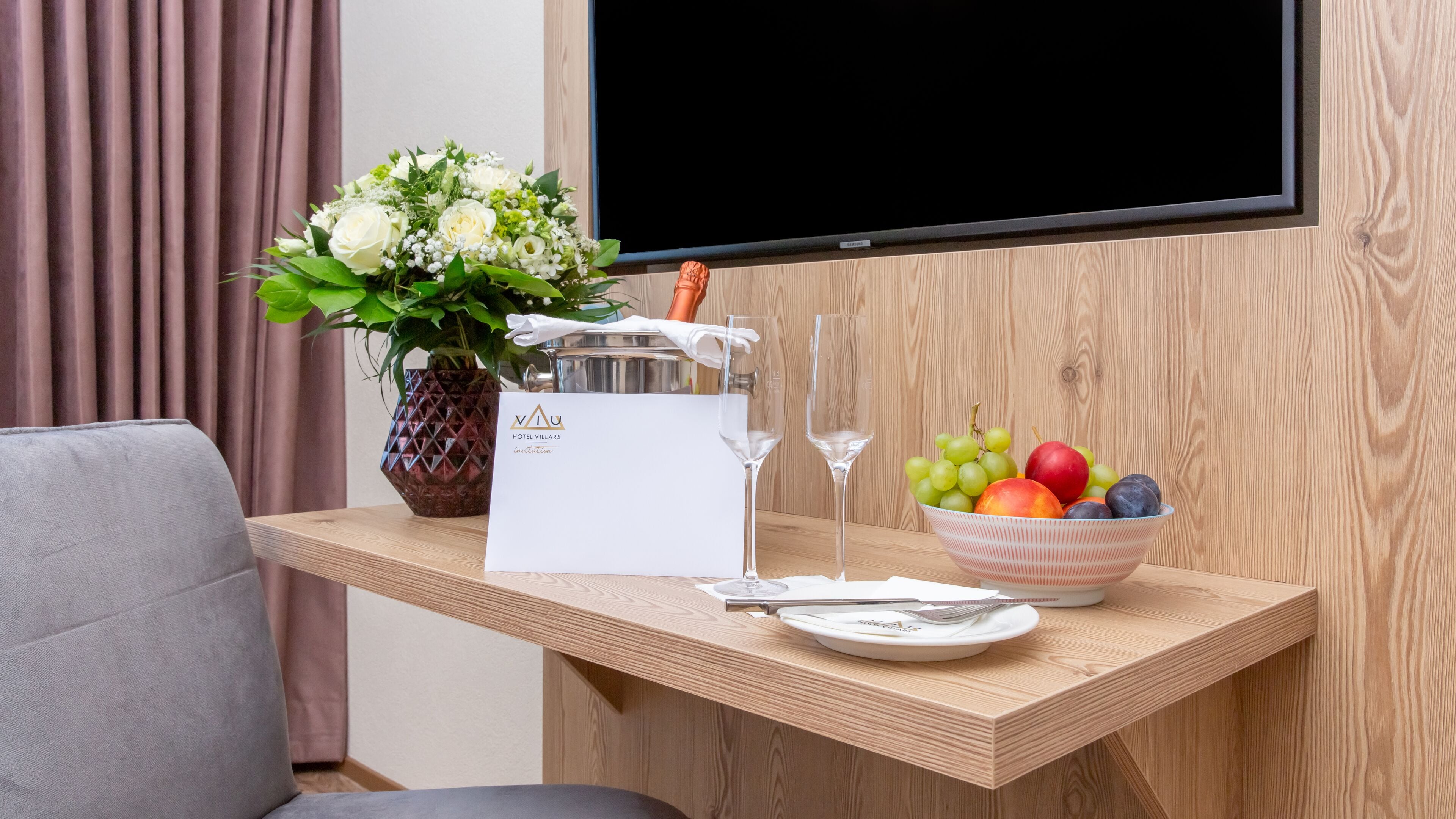A hotel room with a wooden desk, a vase of flowers, a fruit bowl, a wine glass, and a TV mounted on the wall.