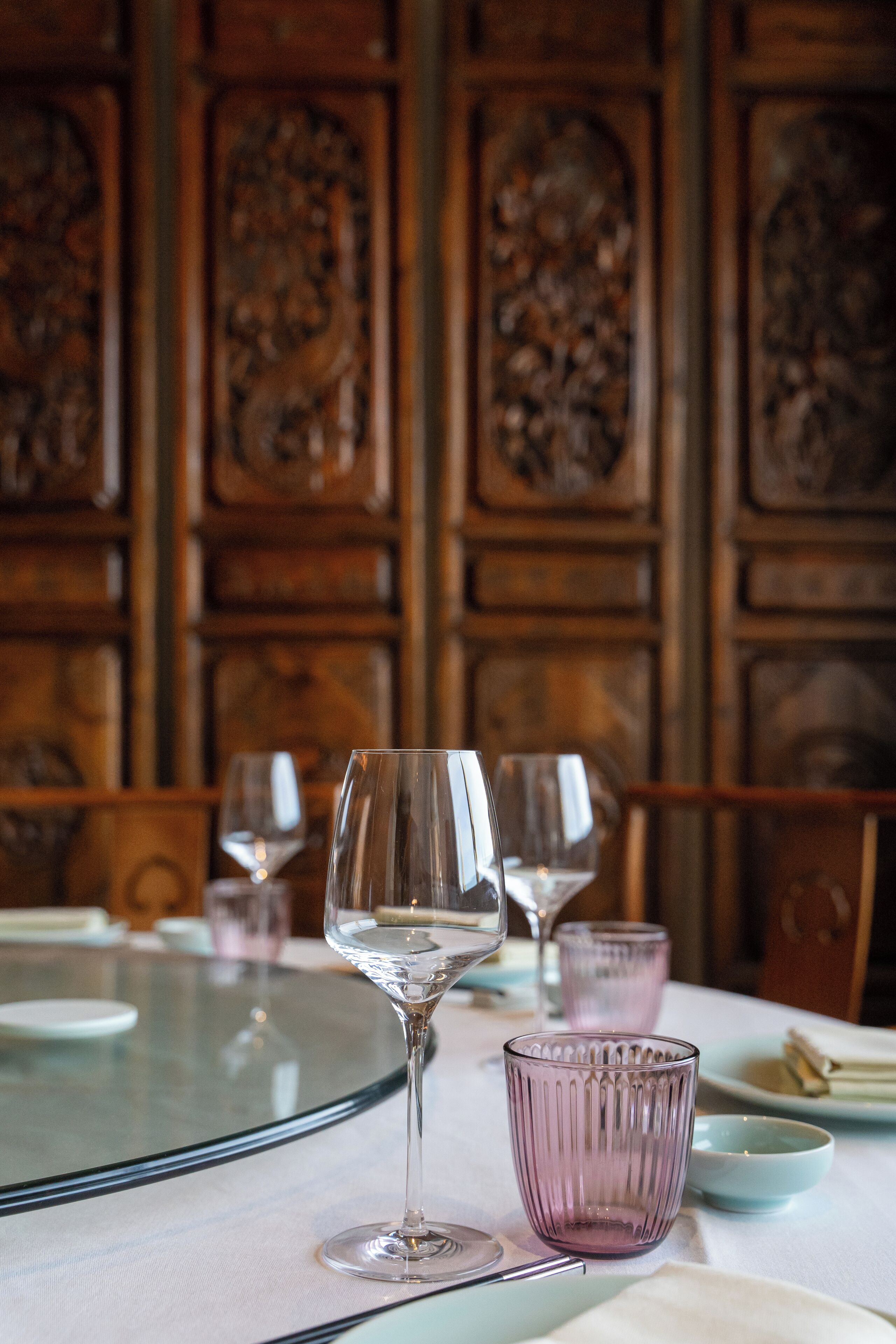 A table with wine glasses and a glass bowl, set against a wooden wall with intricate carvings.