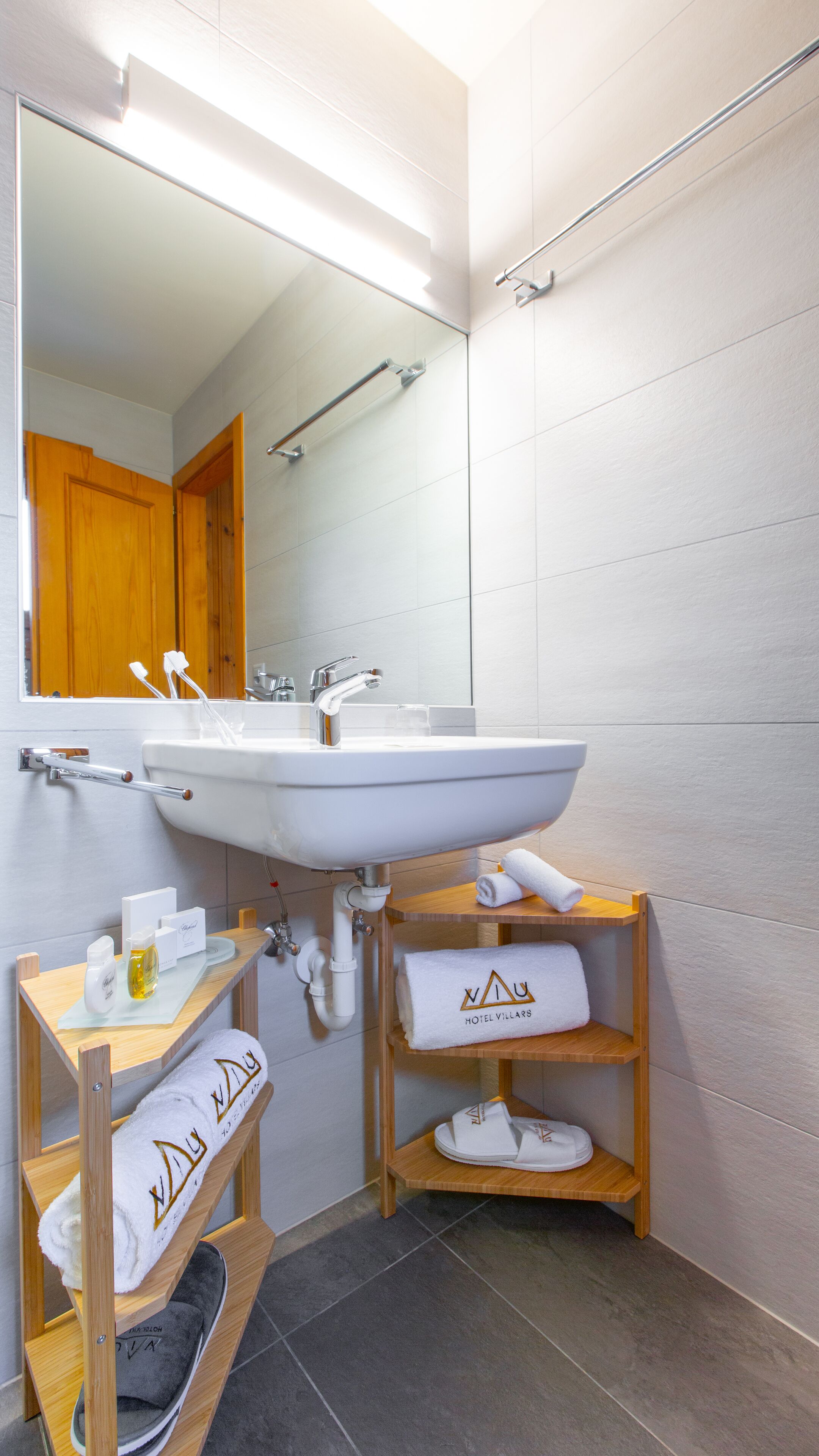 A modern bathroom with a white sink, mirror, and wooden shelves holding towels and slippers.