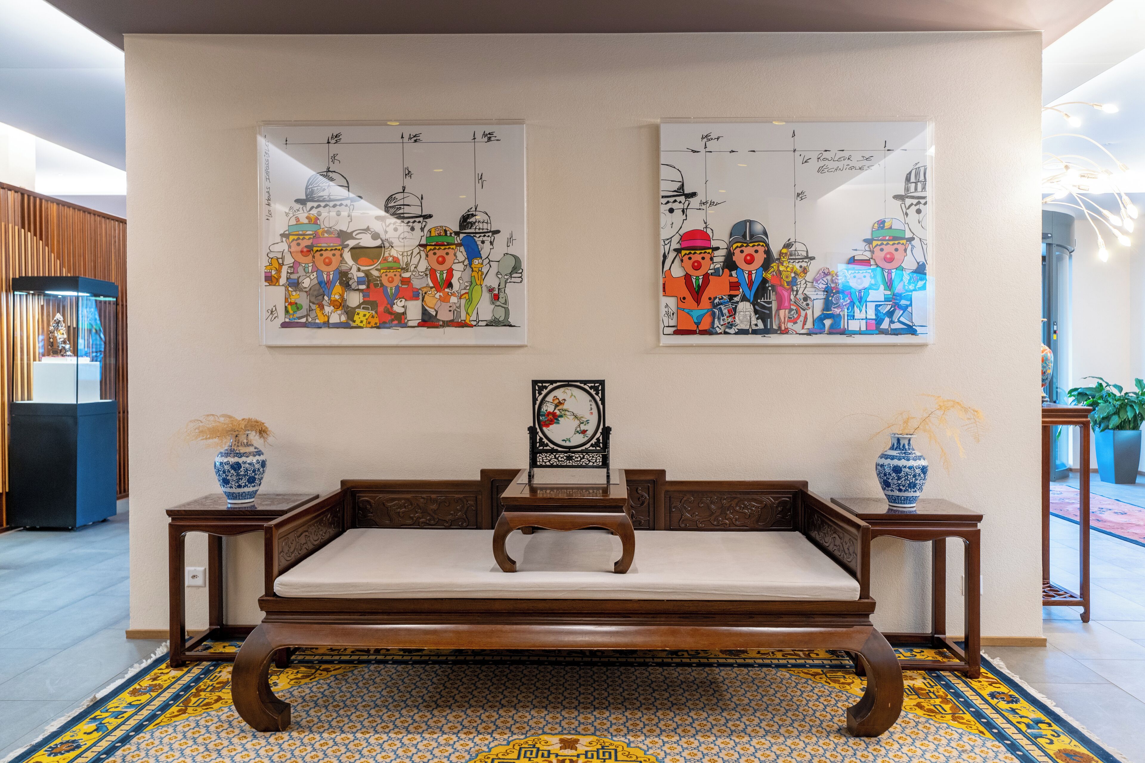 Un sofá de estilo tradicional con un cojín, flanqueado por dos mesitas de madera, cada una con un jarrón azul y blanco. Sobre el sofá, dos piezas de arte mural coloridas muestran personajes de caricatura con vestimenta tradicional.