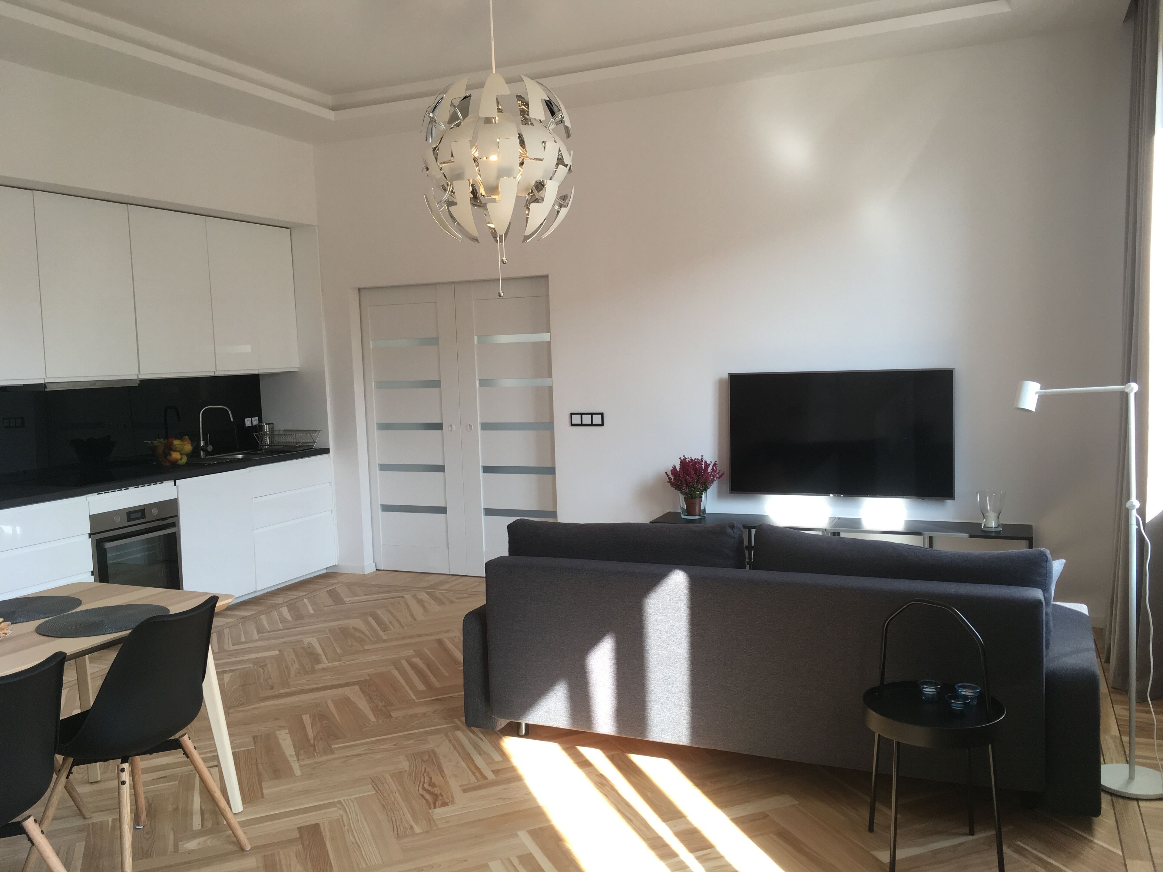 Eine moderne Küche mit weißen Schränken, einer schwarzen Arbeitsplatte und einem eingebauten Backofen. Es gibt ein graues Sofa, einen kleinen Tisch mit Stühlen und einen flachbildschirm-Fernseher an der Wand.