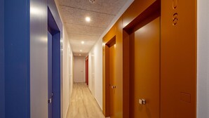 Un pasillo con suelo de madera, puertas azules y anaranjadas, y un techo con iluminación empotrada.