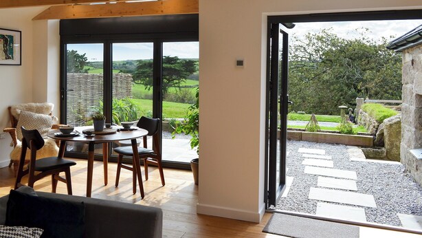 A dining area with a table and chairs, a sofa, and a view of the outdoors through large windows. - The Hen House-UK12928 (Nancherrow, near Penzance)