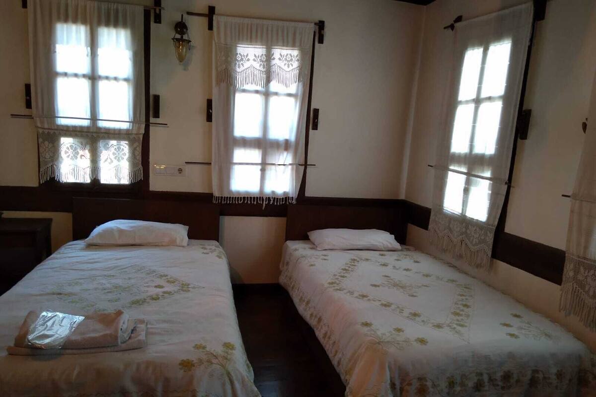 A room with two single beds, each with a white bedspread and a pillow. There are windows with curtains, a wall-mounted lamp, and a wooden headboard.