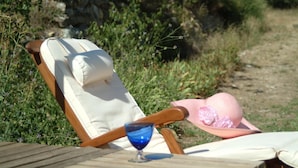 Un fauteuil de jardin en bois avec un coussin, un chapeau rose orné de fleurs et un verre bleu posé sur une table en bois.