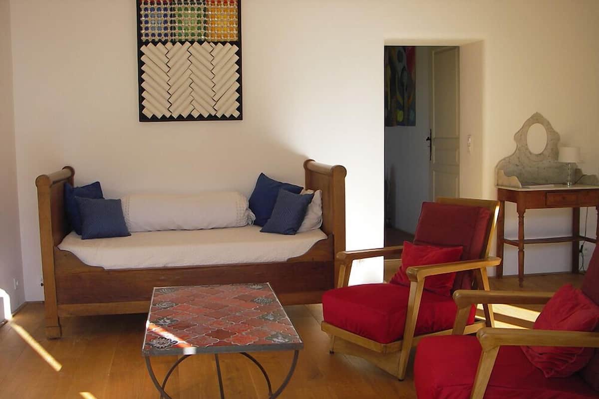 Un salon chaleureux avec un canapé en bois, des fauteuils rouges, une petite table et une œuvre d’art abstraite et colorée accrochée au mur.