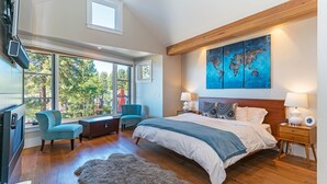 A modern bedroom with a large bed, a world map artwork, a desk, and a view of trees through large windows.