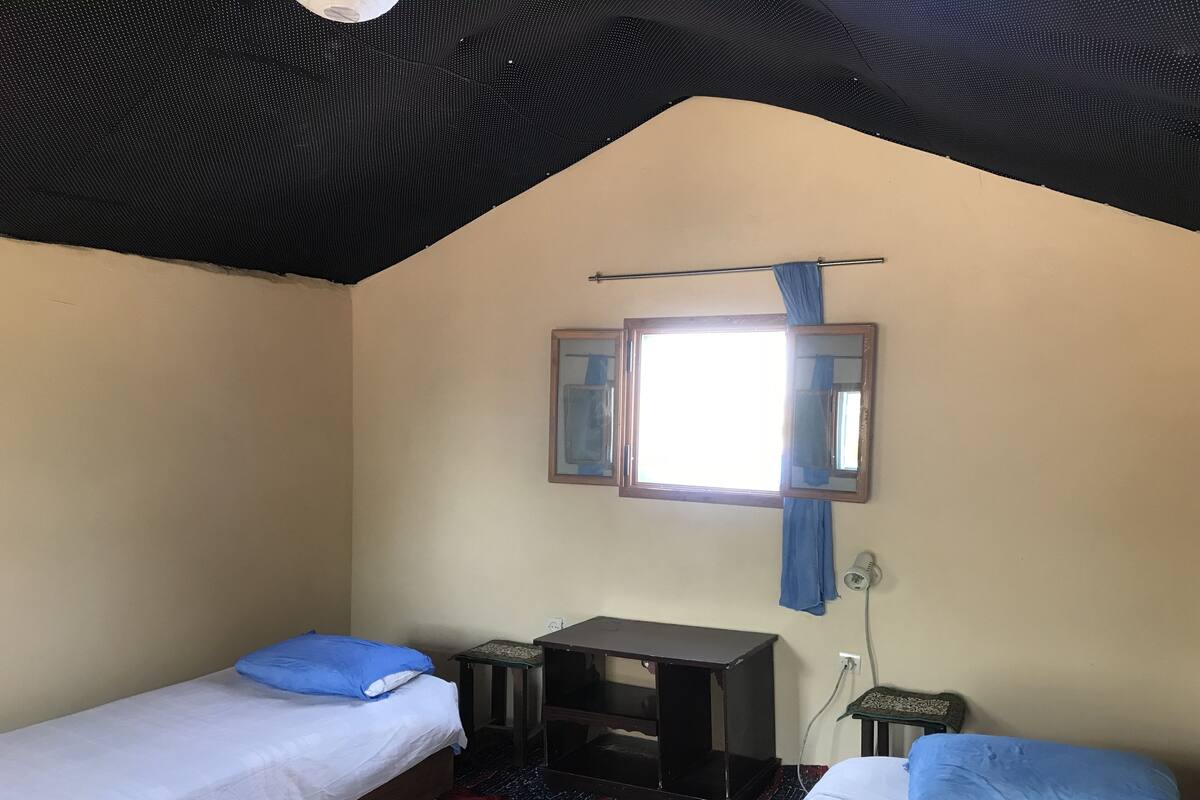 A small, simple room with two single beds, a small table, a mirror, and a window with curtains.