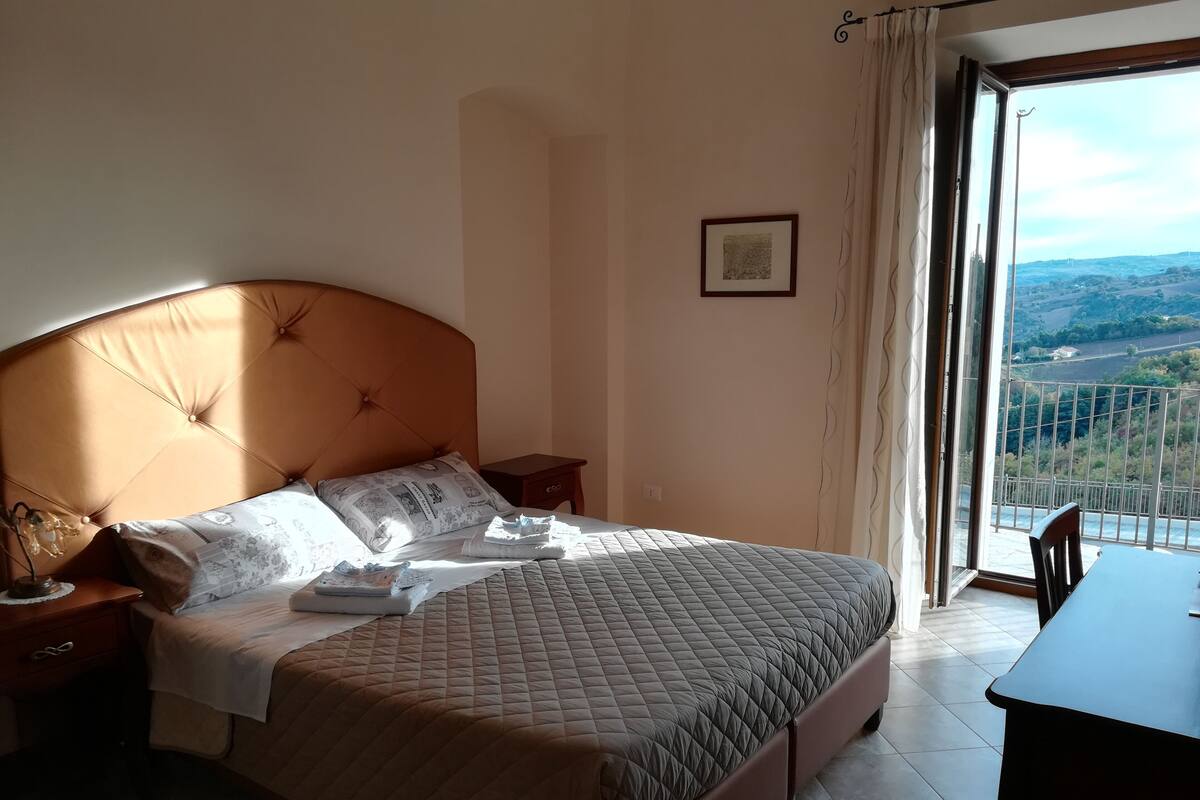 A bedroom with a large bed, a bedside table, a small table, and a view of the outdoors through a window.