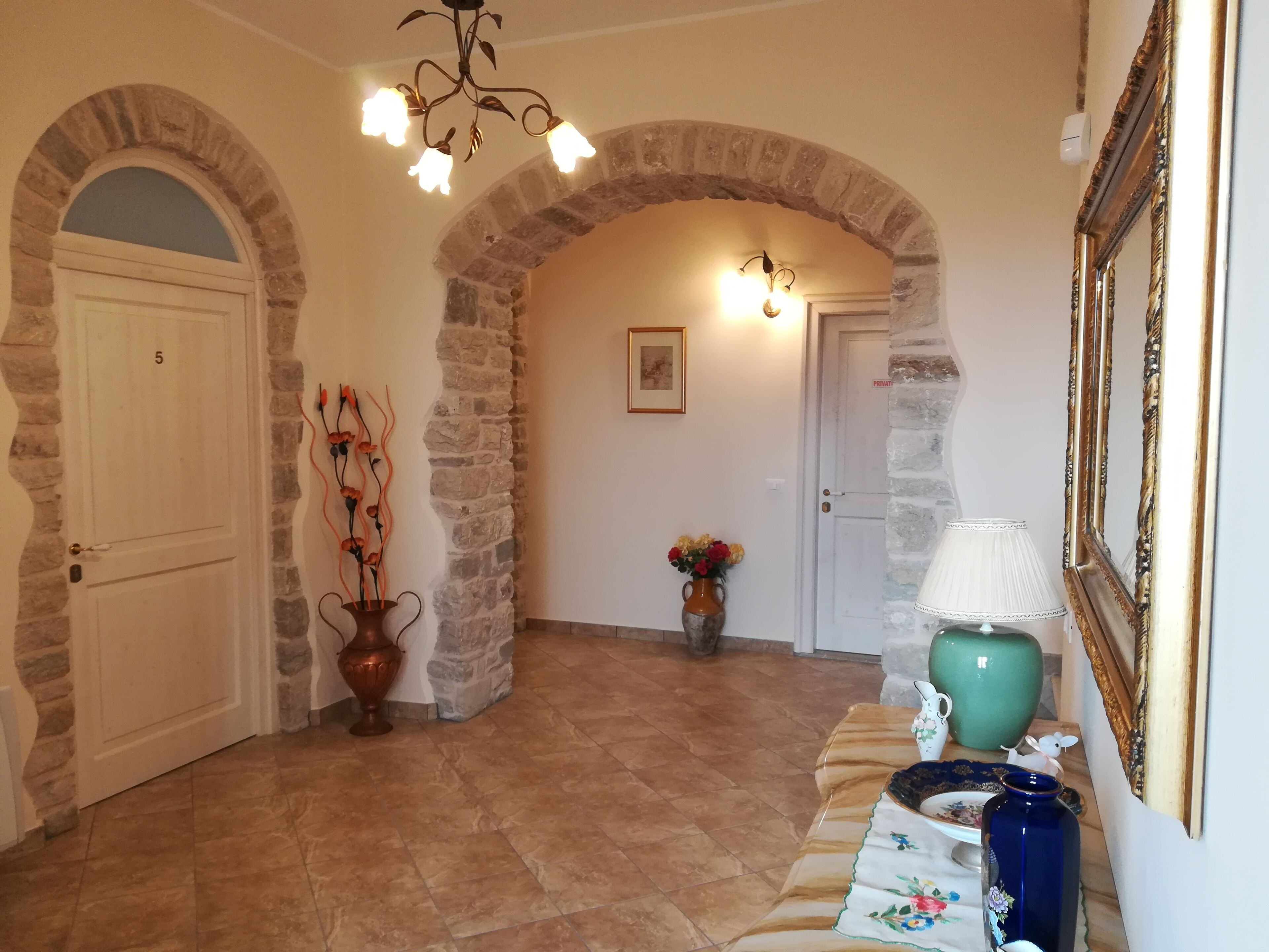 A hallway with stone arches, a door labeled '5', a vase with flowers, and a table with a lamp and decorative items.