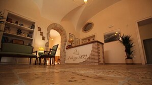 A cozy interior with a stone archway, a reception desk labeled "Palazzo 1892", a green sofa, and a potted plant. - Palazzo 1892 (Castelvetere in Val Fortore)
