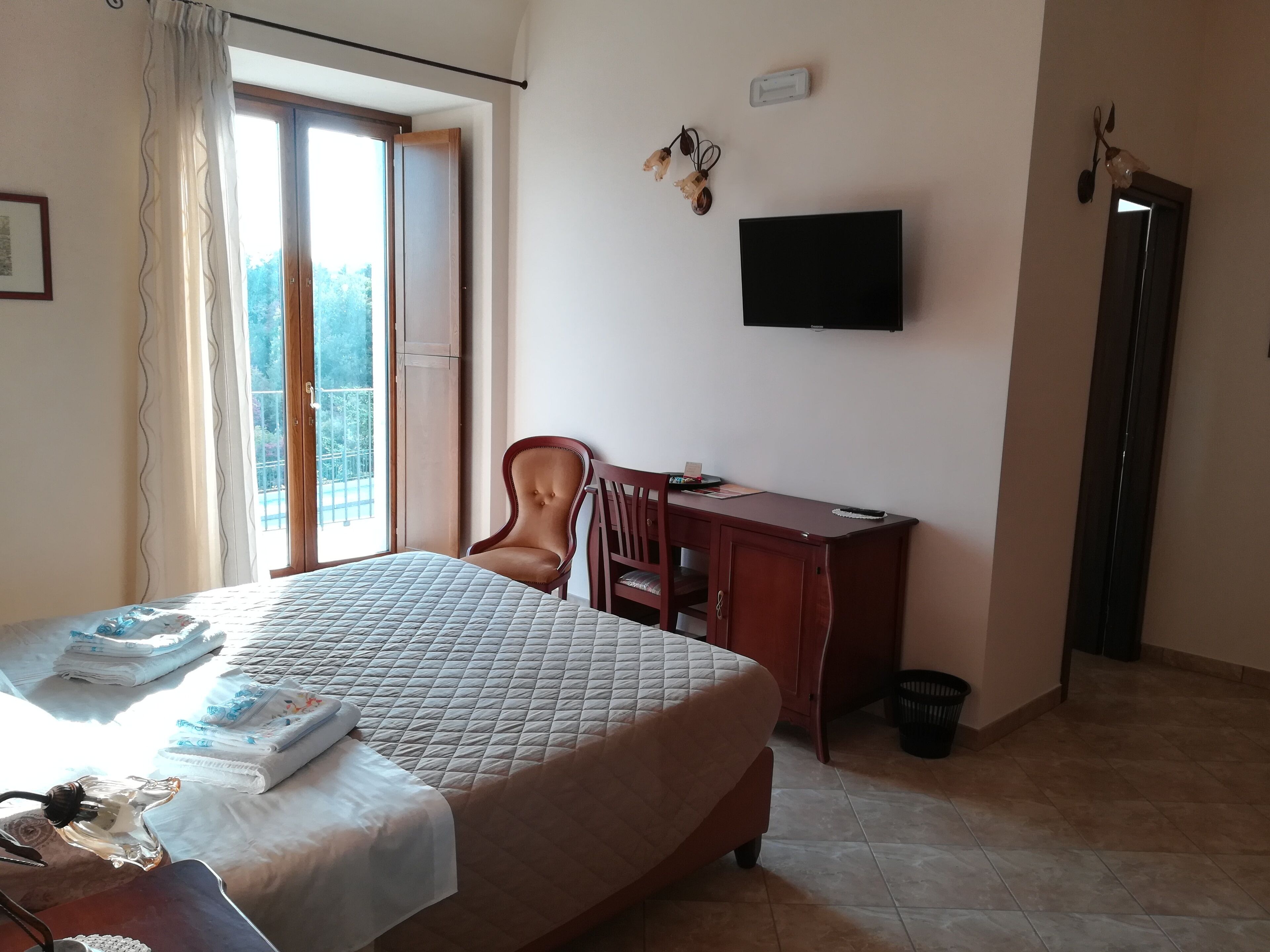 Habitación de hotel con cama, escritorio, silla, televisor y balcón con vista a árboles.