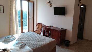 Habitación de hotel con cama, escritorio, silla, televisor y balcón con vista a árboles.