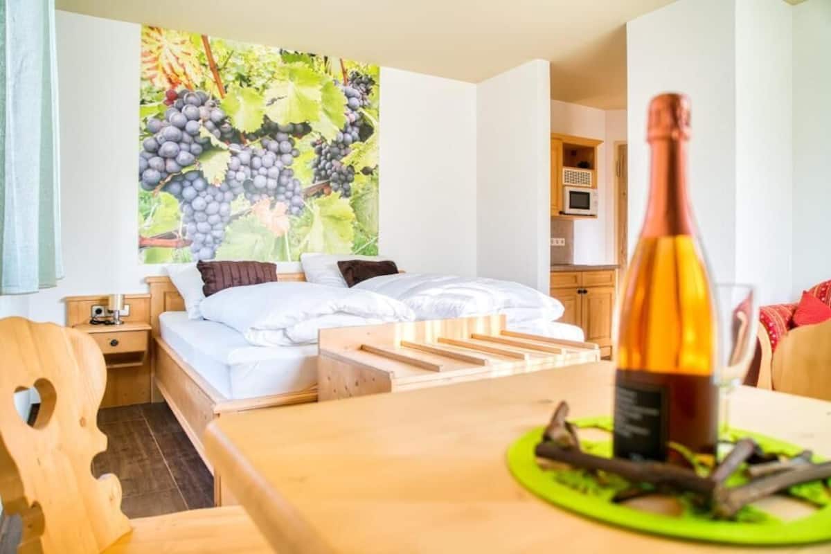 A bedroom with a bed, a wooden nightstand, a green table with a bottle and branches, and a kitchen in the background.