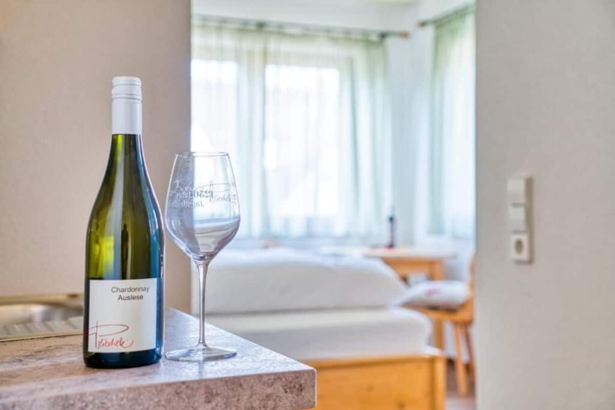 A bottle of Chardonnay Auslese and a wine glass on a marble countertop in a hotel room with a bed and a window with curtains.