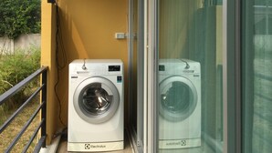 A balcony with two washing machines, an air conditioning unit, and a glass door leading to a balcony with a view of greenery.