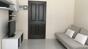 A living room with a dark wooden door, a white entertainment unit with a television, and a grey sofa with white cushions.