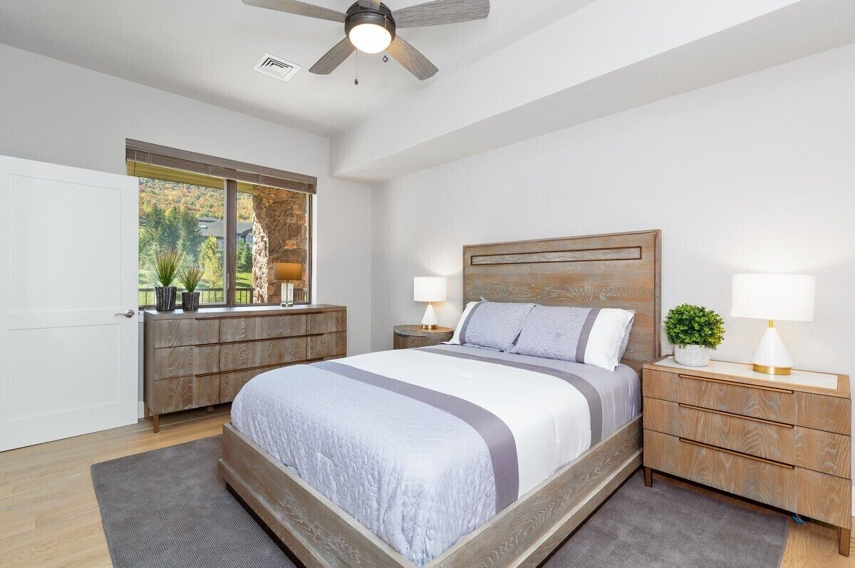 A bedroom with a large bed, wooden headboard, two bedside tables with lamps, a window with a view of trees, and a ceiling fan.
