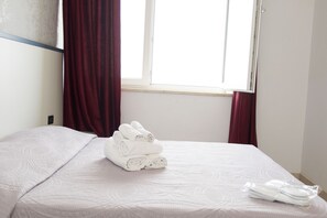 A neatly made bed with a white comforter and rolled towels, a window with red curtains, and a plain wall.