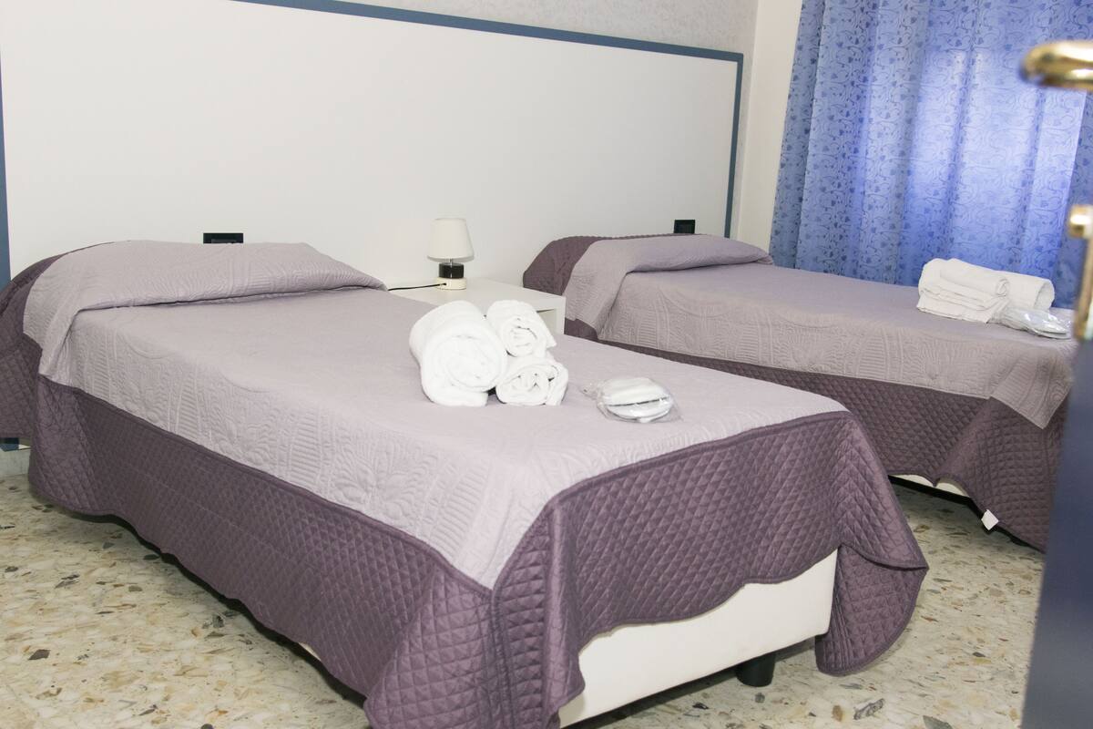 Habitación de hotel con dos camas, cada una con cubrecama morado y almohadas blancas. Hay toallas y una mesita con una lámpara.