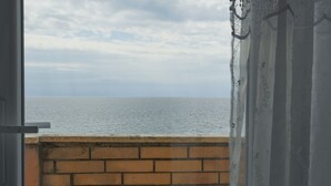 Vista a través de una ventana con un muro de ladrillo y una cortina, con vista al mar tranquilo bajo un cielo nublado.