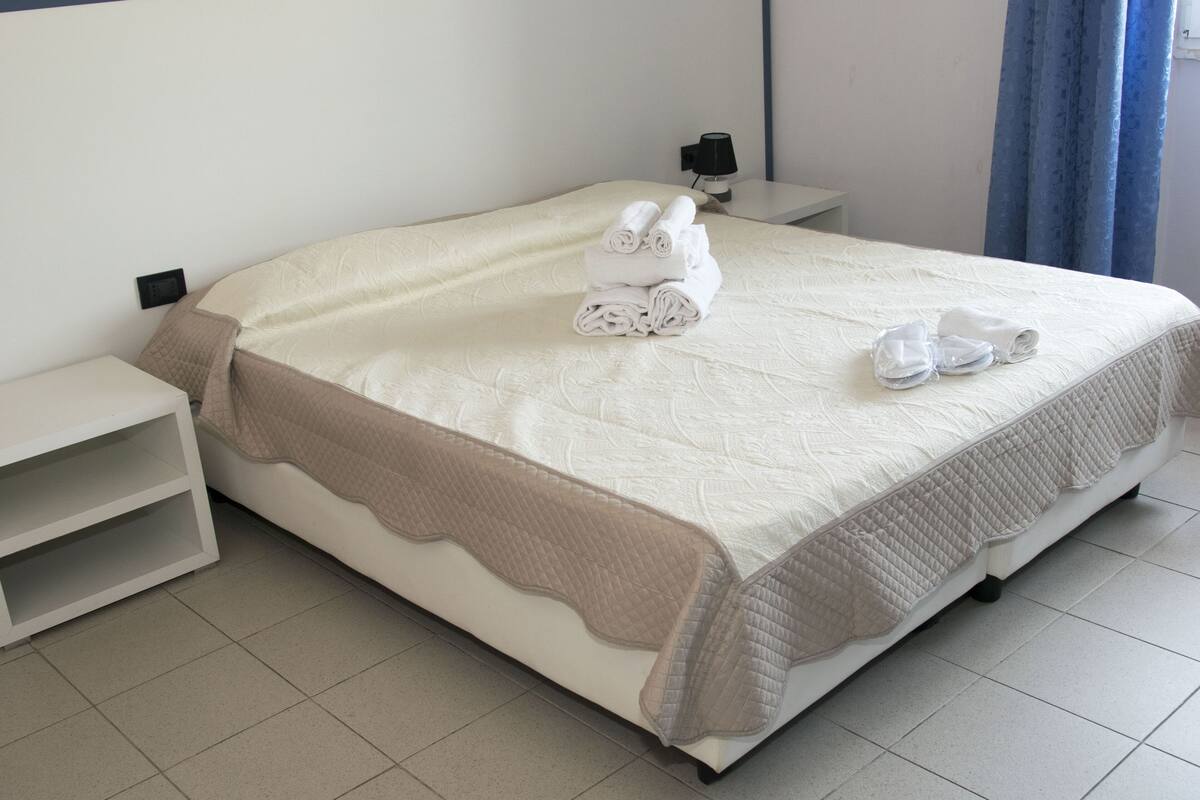 Una cama bien tendida con un edredón acolchado y una manta beige. Hay dos toallas enrolladas sobre la cama.