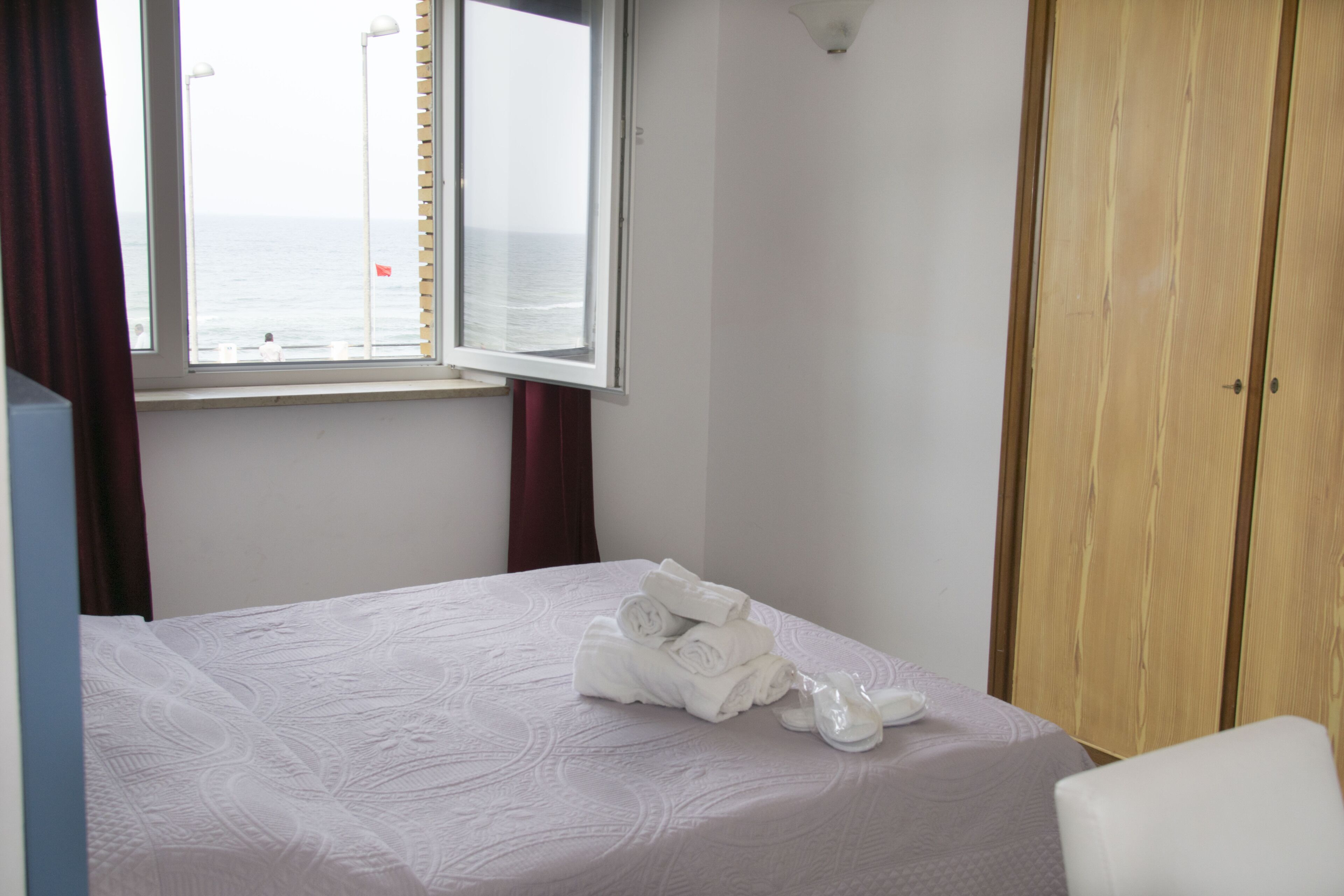 A hotel room with a bed, a window with a view of the sea, and a wooden wardrobe.
