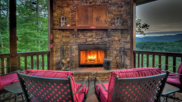 A stone fireplace with a fire, surrounded by red cushioned chairs on a deck with a view of a forest and mountains. - Leatherwood Lodge- Breathtaking Views | Hot Tub | Outdoor Fireplace (Cherry Log)