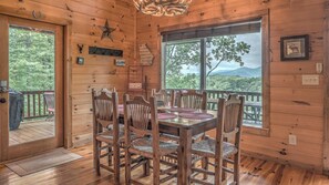 A wooden cabin interior with a dining table, six chairs, a star decoration, a coat rack, and a window with a view of trees and mountains. - Leatherwood Lodge- Breathtaking Views | Hot Tub | Outdoor Fireplace (Cherry Log)