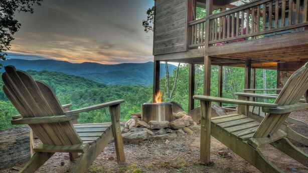 Eine Holzhütte mit zwei Adirondack-Stühlen, die auf eine Feuerstelle ausgerichtet sind, mit Blick auf eine gebirgige Landschaft.