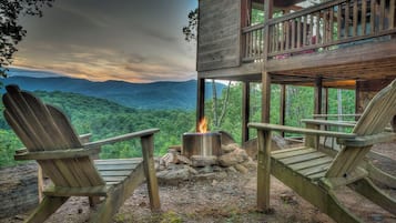 A cabin with two Adirondack chairs facing a fire pit, overlooking a mountainous landscape.