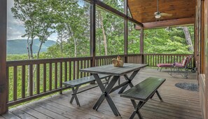A covered outdoor deck with wooden benches and a picnic table, surrounded by a wooden railing, overlooking a lush green landscape with trees and mountains. - Leatherwood Lodge- Breathtaking Views | Hot Tub | Outdoor Fireplace (Cherry Log)