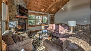 A cozy living room with a stone fireplace, wooden ceiling, and large windows.