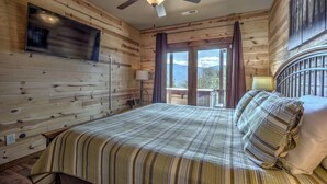 A wooden cabin bedroom with a bed, a TV mounted on the wall, and a balcony with a view of the mountains.