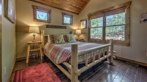 Quarto aconchegante com cama de madeira, duas luminárias de apoio, um tapete vermelho e uma janela grande com vista para árvores.