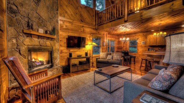 Un acogedor interior de cabaña con chimenea de piedra, paredes de madera, sala de estar con sofá y mesa de centro, zona de cocina y un balcón con barandilla.