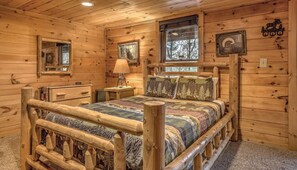 Un dormitorio de cabaña de madera con una cama de madera, una mesita de noche, una lámpara y una ventana con vistas a árboles.