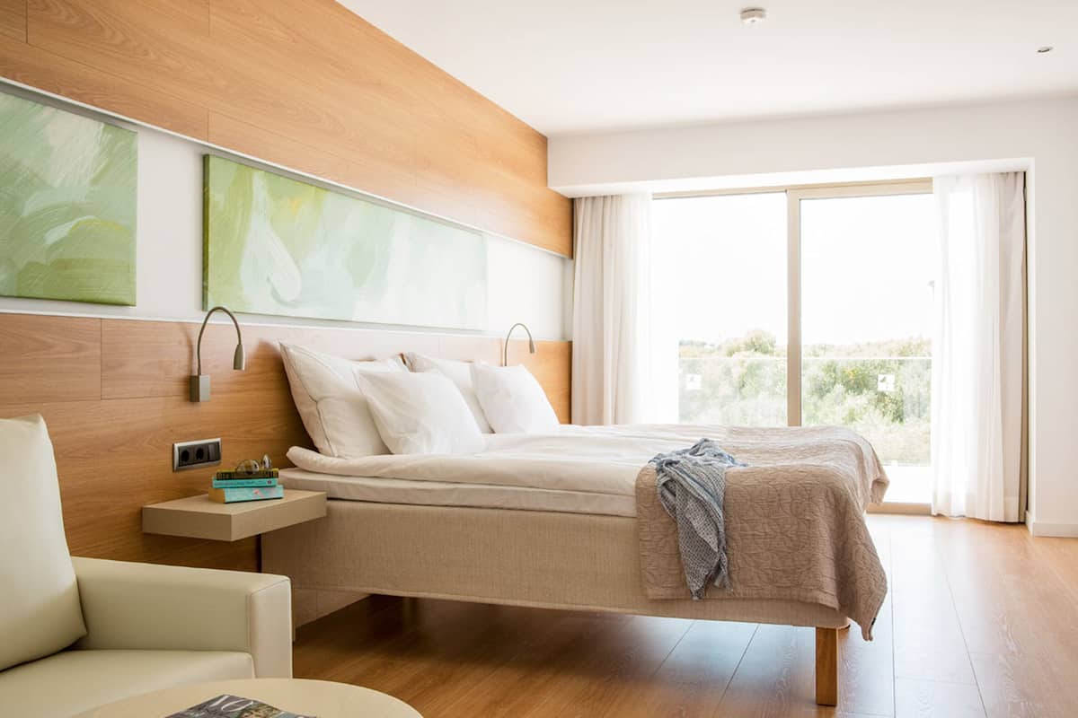 A modern bedroom with a large bed, a nightstand, a chair, and a view of greenery through the window.