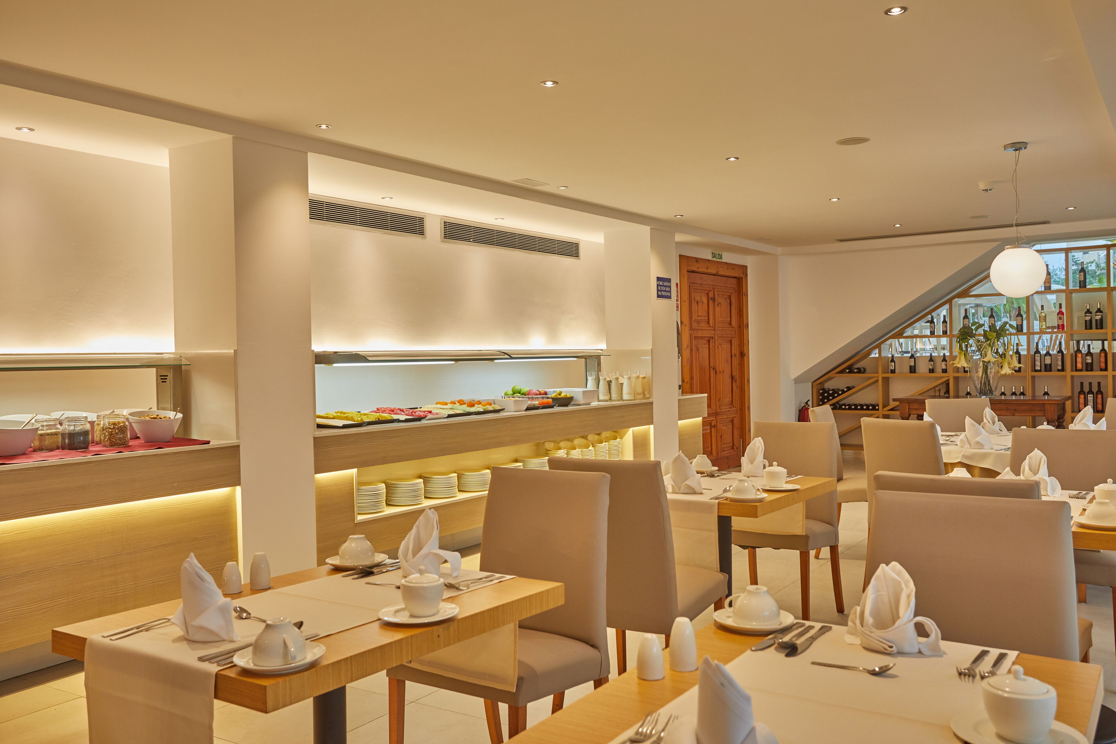 Un espace repas moderne avec des tables dressées pour le petit-déjeuner, un buffet proposant divers plats et, au fond, un présentoir à vins.