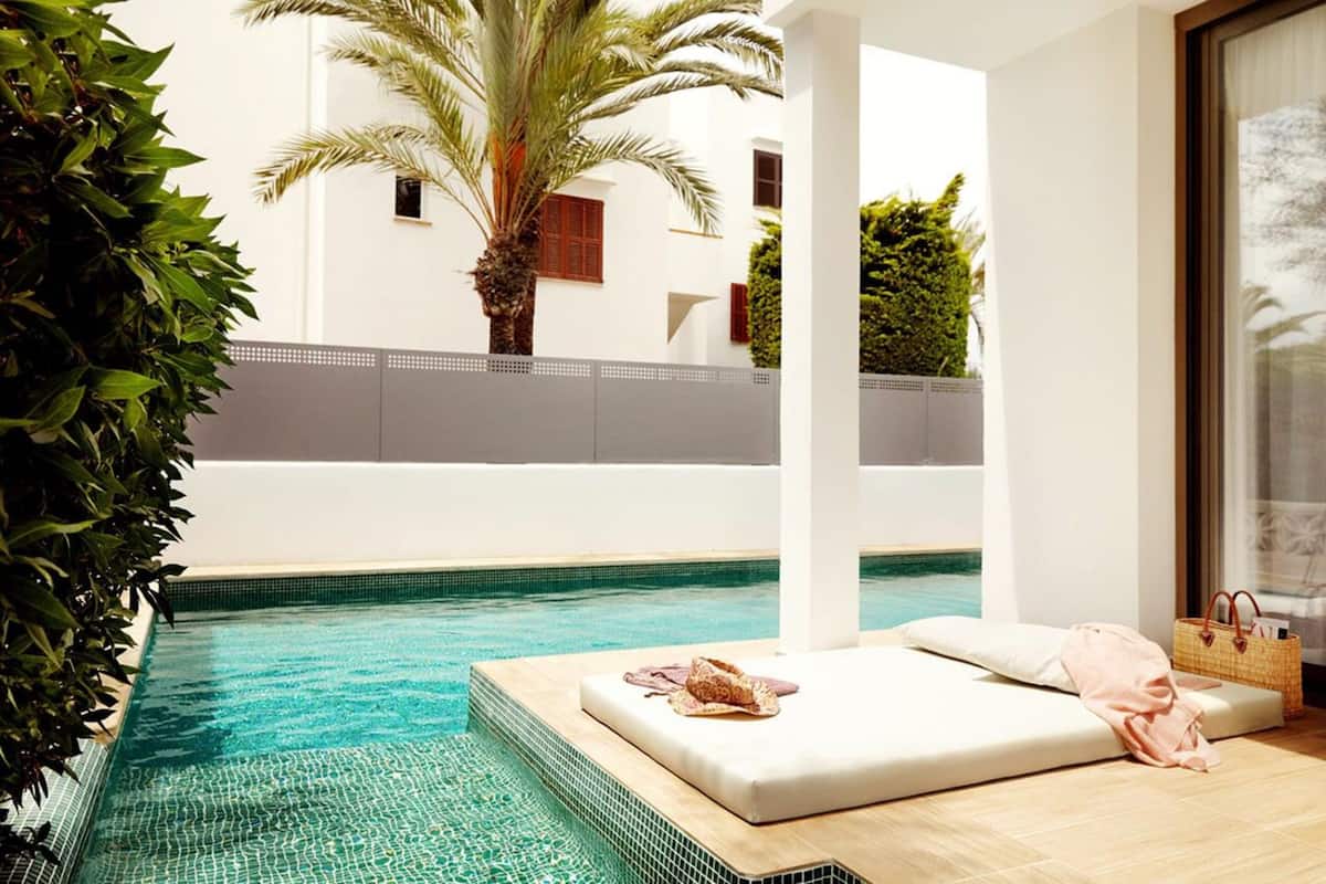 A poolside area with a lounge chair and a small table, surrounded by greenery and a white building in the background.