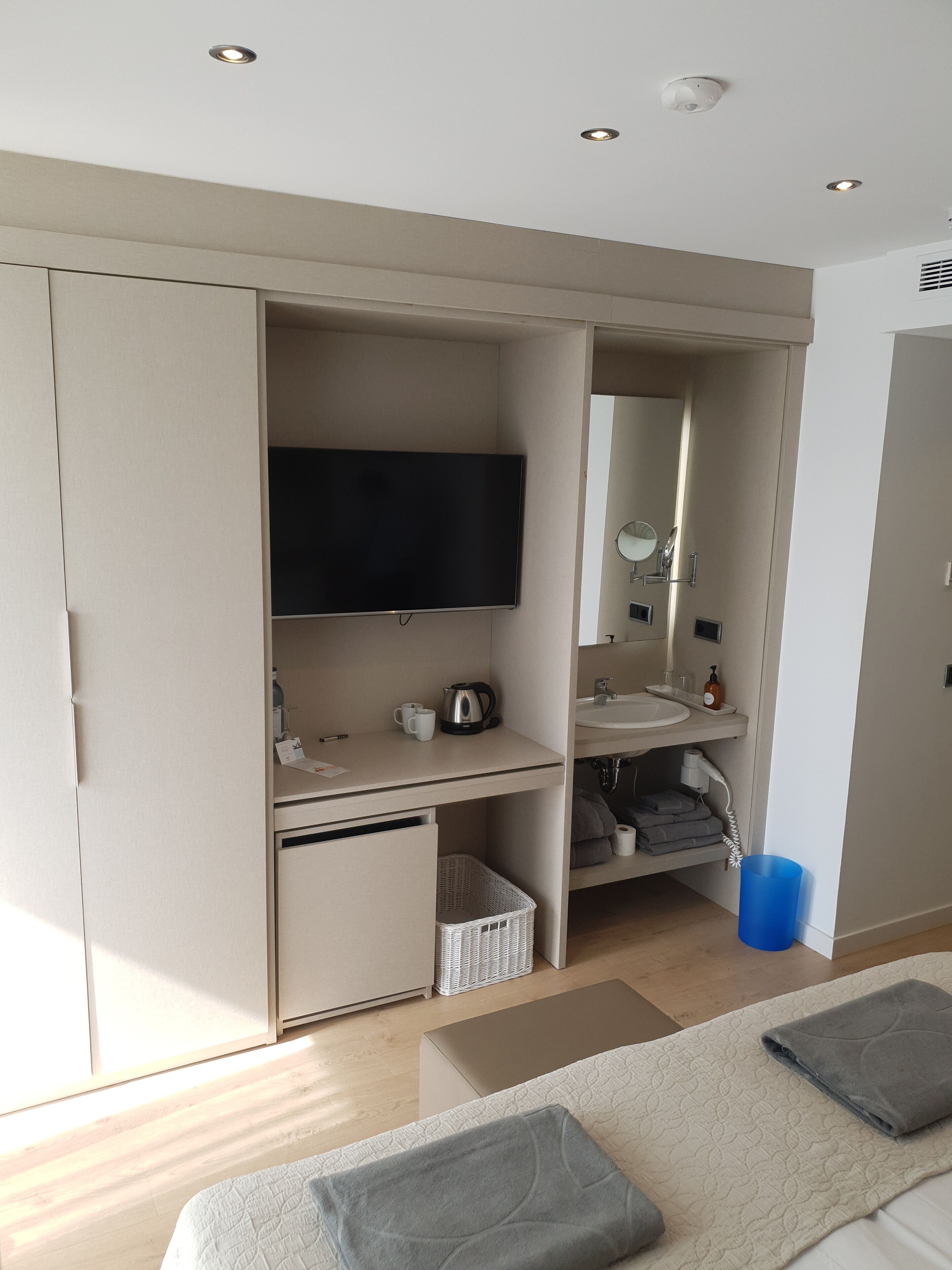 A modern bedroom with a bed, a TV, a desk with a kettle, a sink, and a mirror.