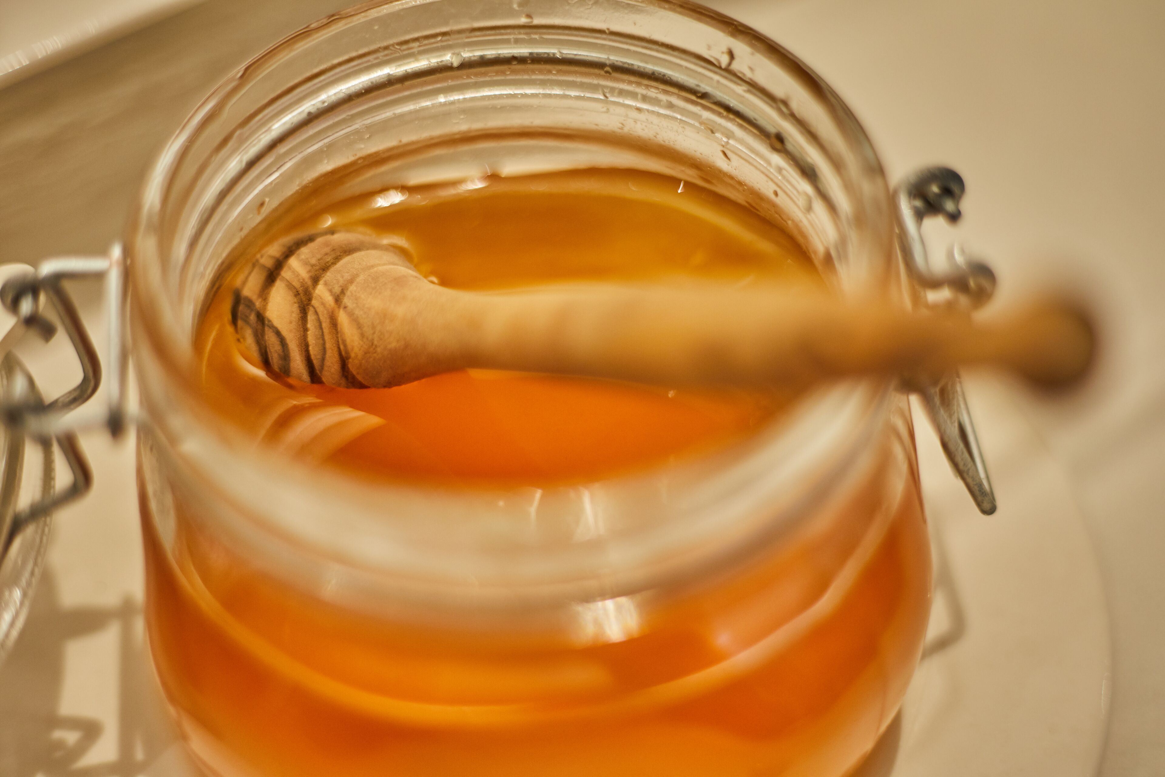 A glass jar with a metal clasp and a wooden honey dipper inside, containing a golden liquid.