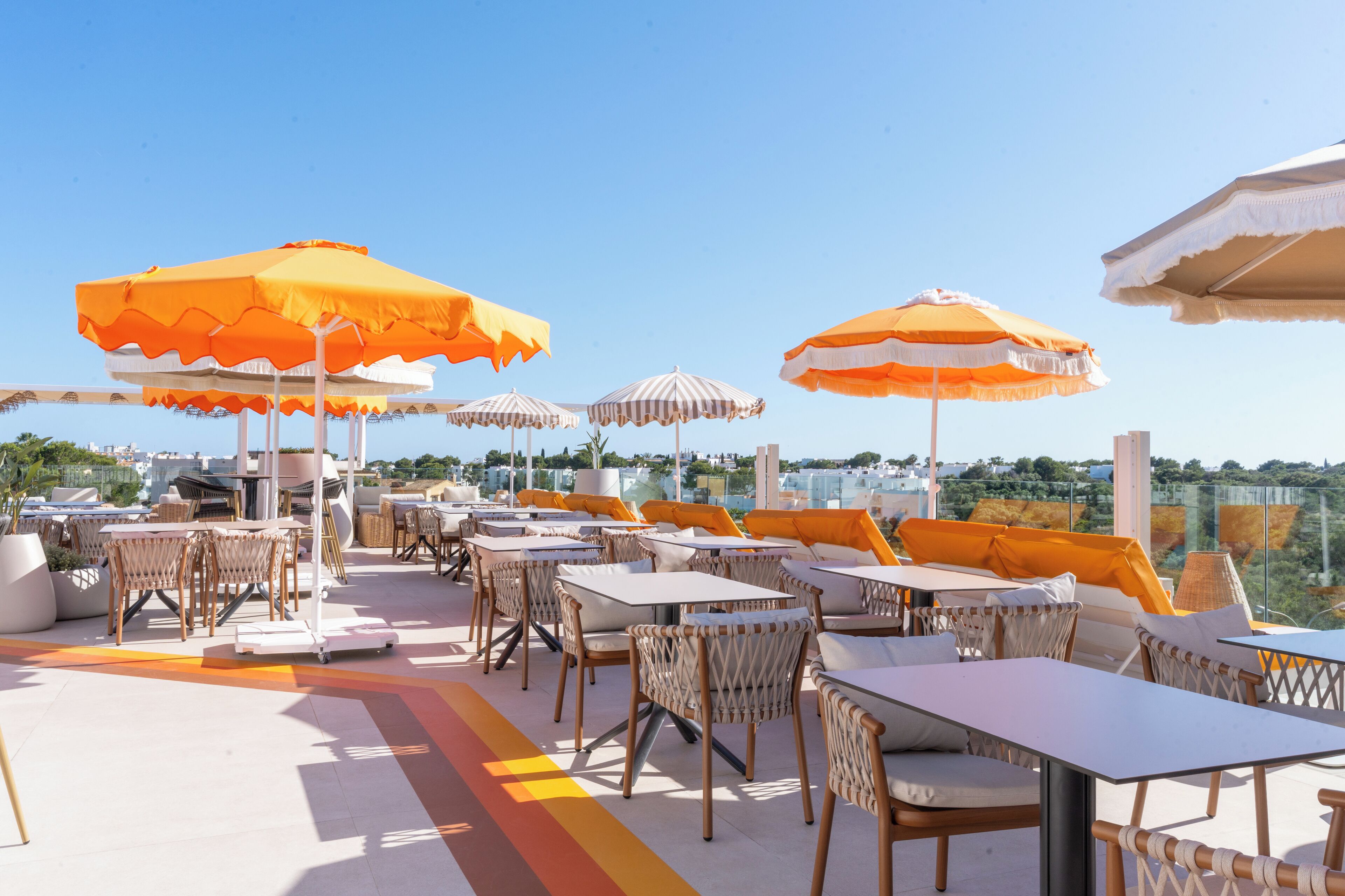 An outdoor seating area with tables and chairs, orange and white striped umbrellas, and a view of the city.