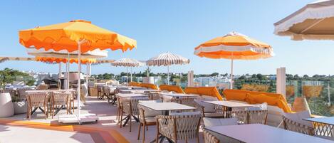 An outdoor seating area with tables and chairs, orange and white striped umbrellas, and a view of the city.